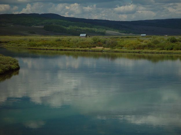 Farm Buildings Behind A Tranquil Green River. Photo by Dave Bell.